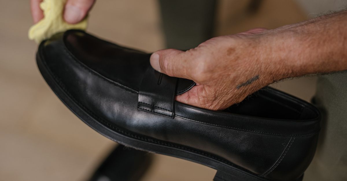 A person's hand polishes a black leather loafer with a yellow cloth