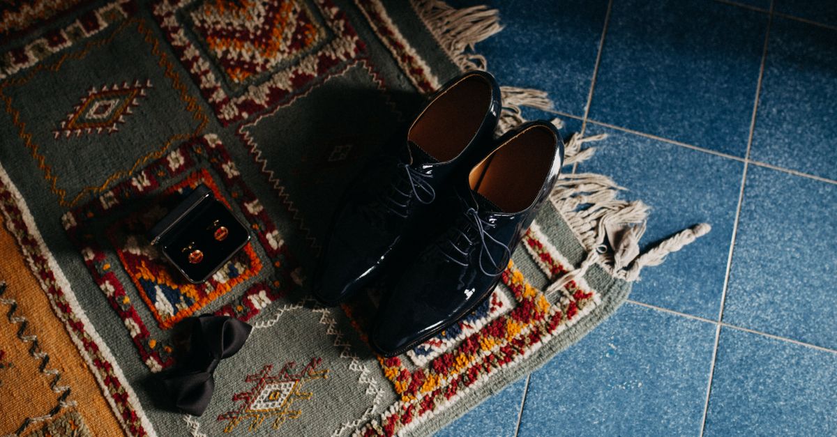 A pair of glossy black patent dress shoes resting on a dark, patterned textile rug alongside cufflinks and a bow tie.