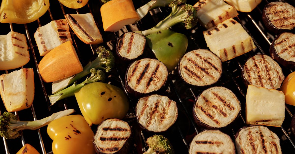 Sliced eggplant rounds with charred grill marks are arranged on a grill grate alongside peppers, squash, and broccoli.