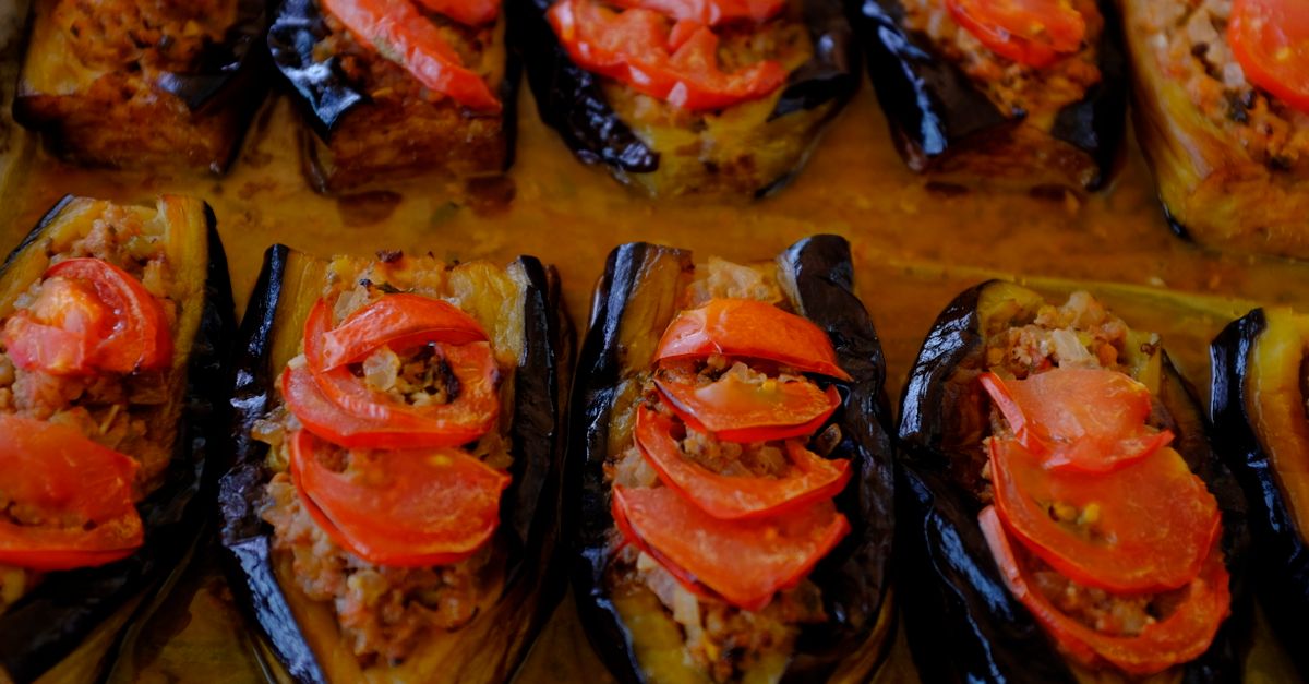 Rows of stuffed eggplant halves topped with sliced tomatoes, baked in a dish with rich sauce.