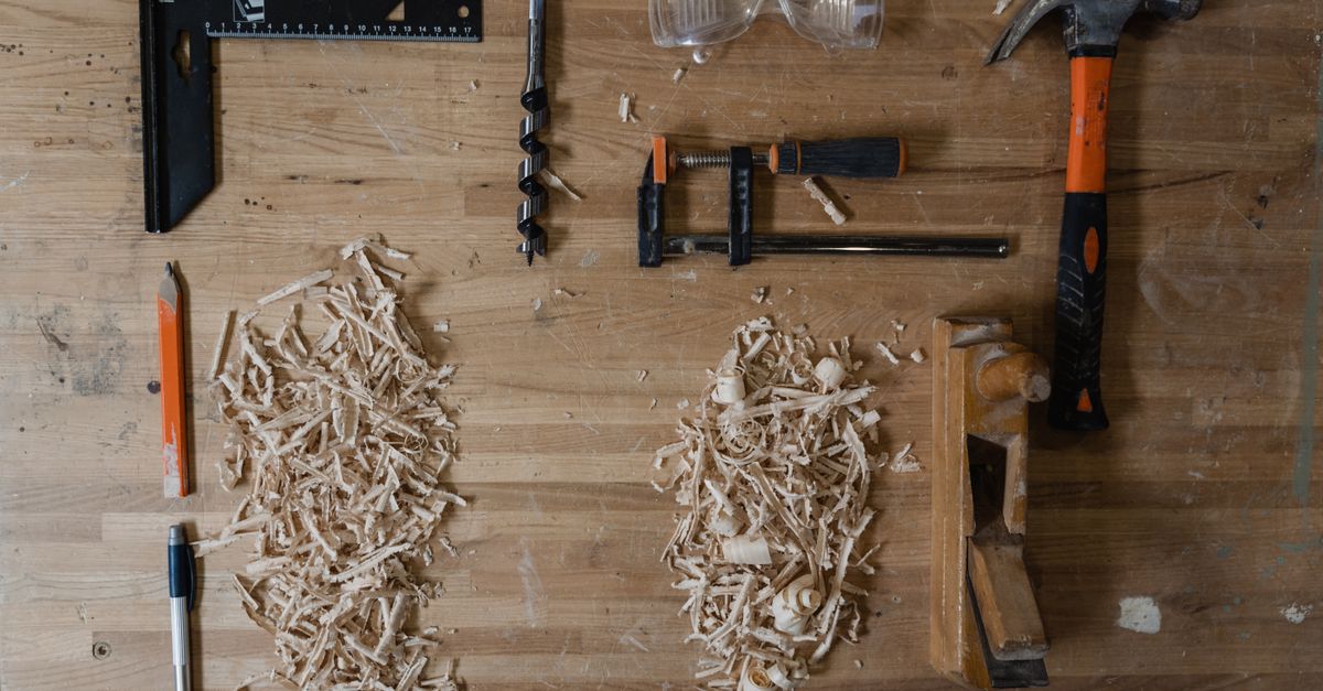 A wooden workbench viewed from above with a square ruler, drill bit, safety glasses, a clamp, a hammer, wood shavings, and a hand plane arranged neatly.