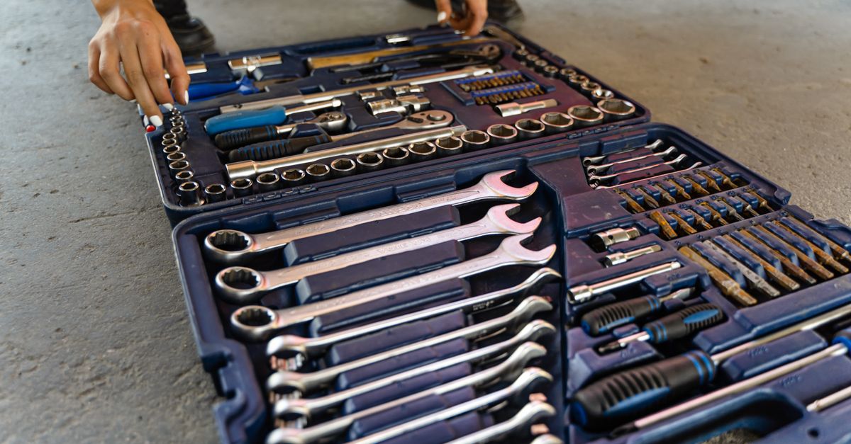 A person reaches into an open tool case laid out on the ground, filled with organized wrenches, sockets, and screwdrivers.
