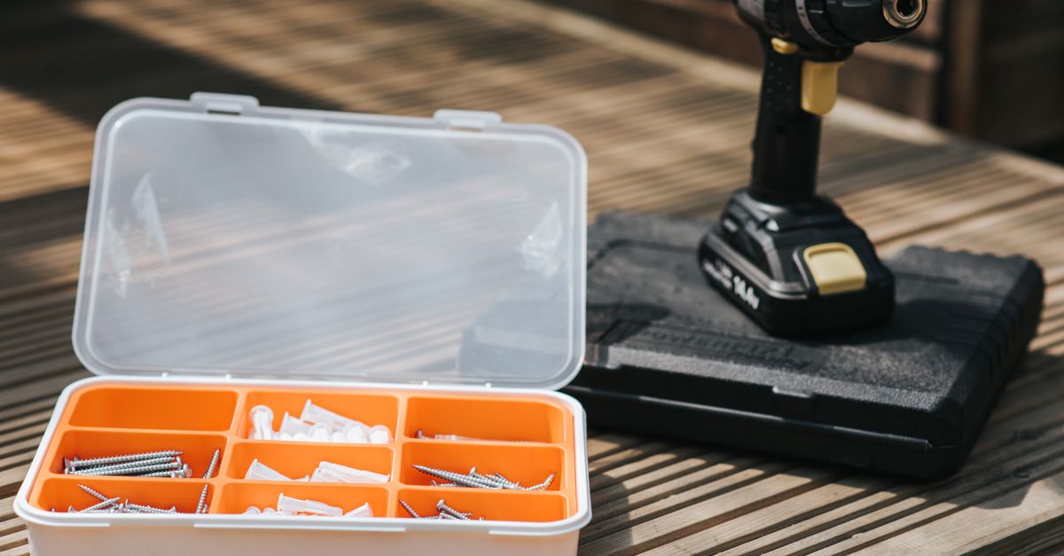 A cordless drill rests on a wooden deck beside an open plastic organizer case filled with screws and wall anchors.