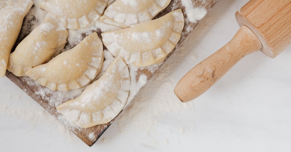 A wooden rolling pin rests beside crimped, flour-dusted dumpling wrappers arranged on a wooden board.