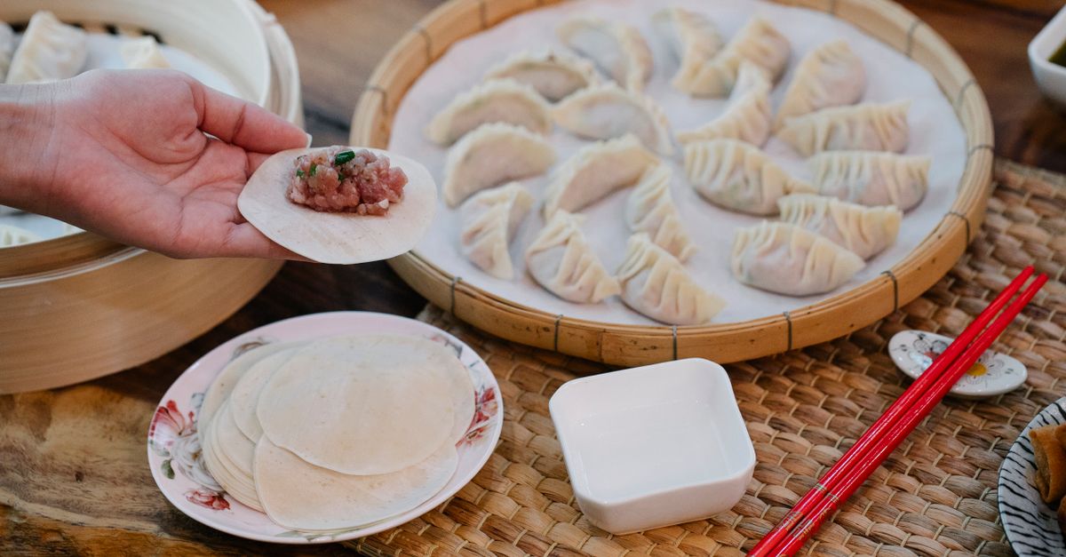 A hand holds a raw dumpling wrapper filled with meat over a plate of flat wrappers, with a bamboo tray of pleated uncooked dumplings and a small dish in the background.