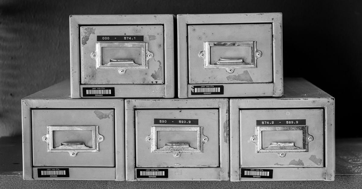 A stack of five worn metal card catalog storage boxes with label holders, stacked two on top of three on a shelf.