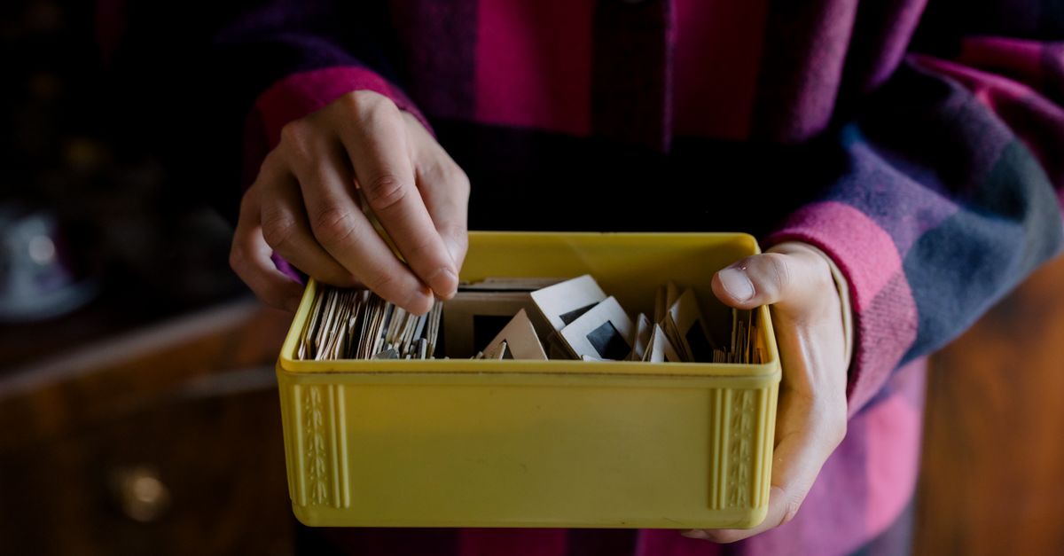 A person's hands sorting through a yellow plastic box filled with photograph slides and prints.