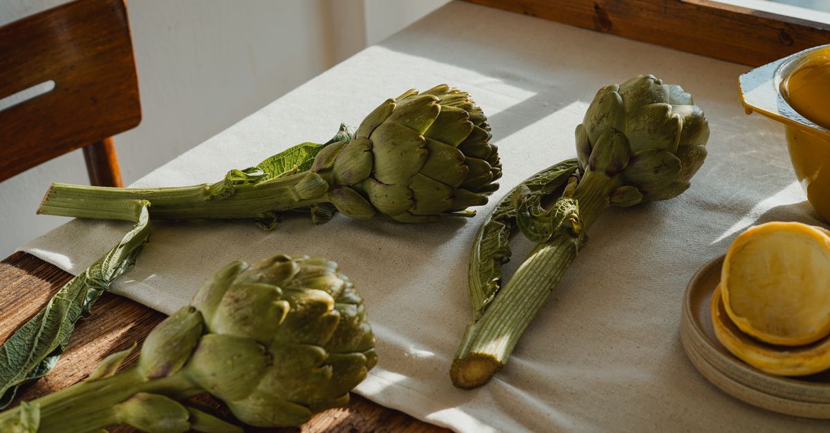 Whole raw artichokes with long stems lying on a cloth-covered table beside stacked lemon slices and a yellow bowl.