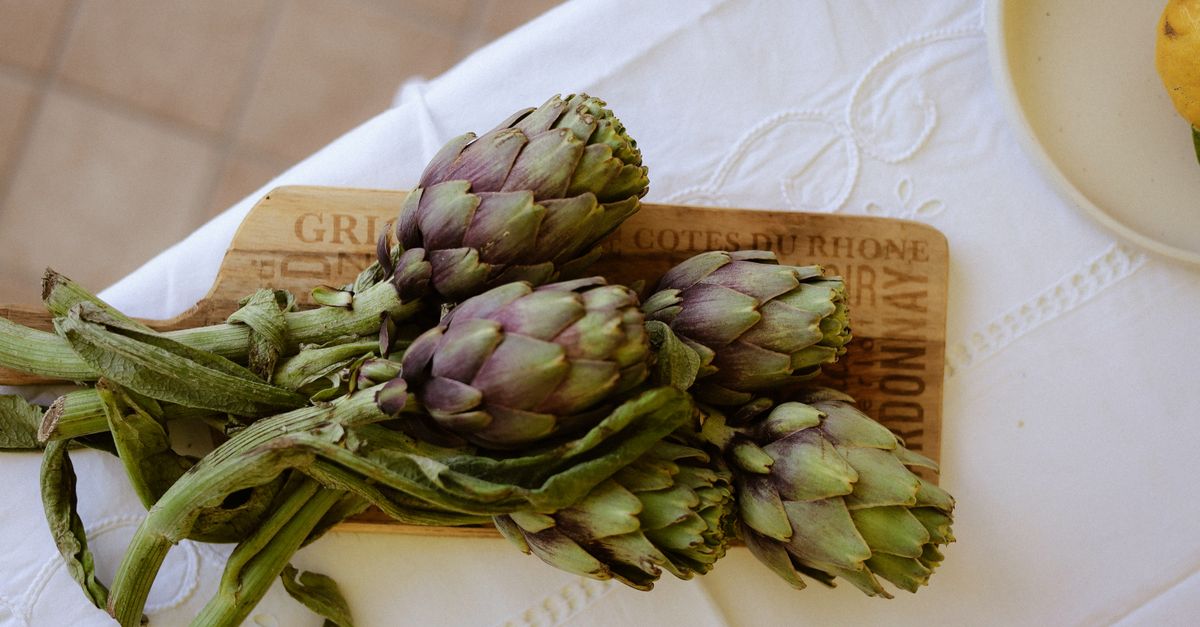Several fresh green artichokes with long stems resting on a wooden cutting board with printed text, set on a white tablecloth.