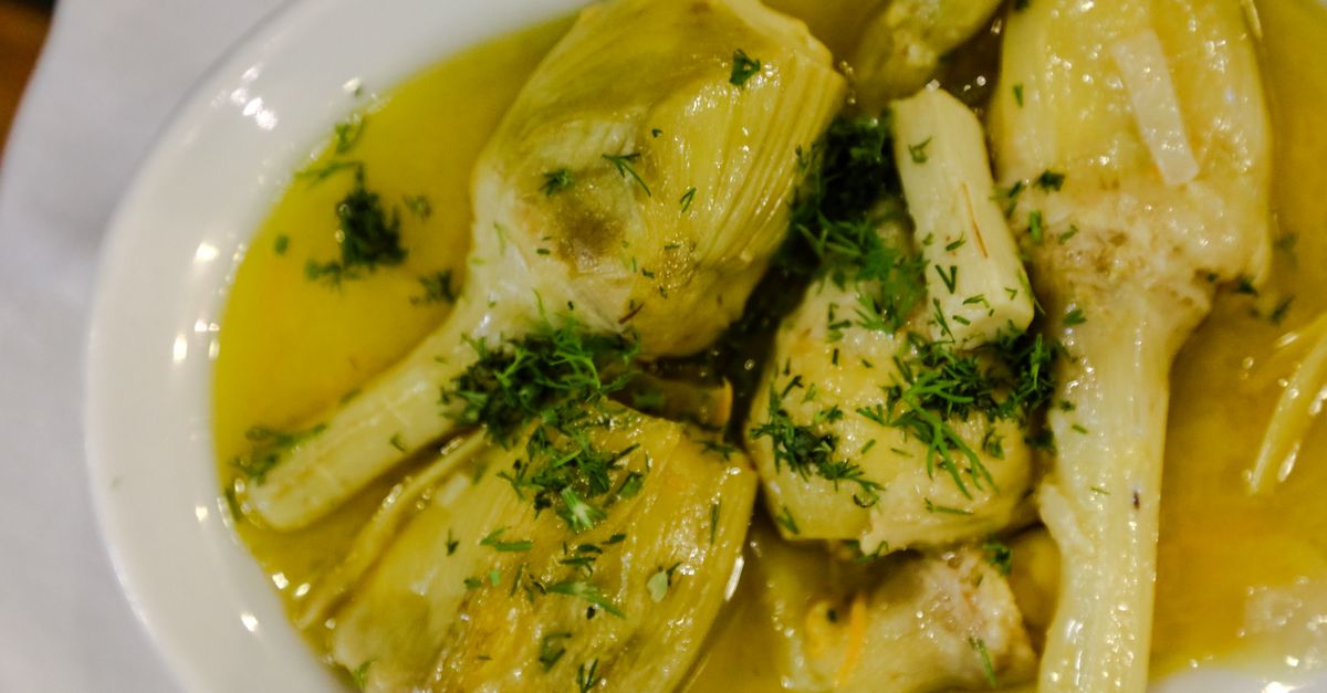 A white bowl of cooked artichoke hearts and stems in a golden olive oil broth garnished with chopped herbs.