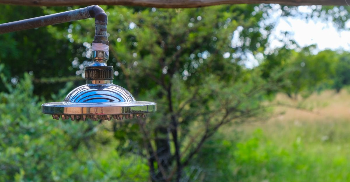 A close-up of a chrome shower head and pipe hanging outdoors against a blurred green natural background.