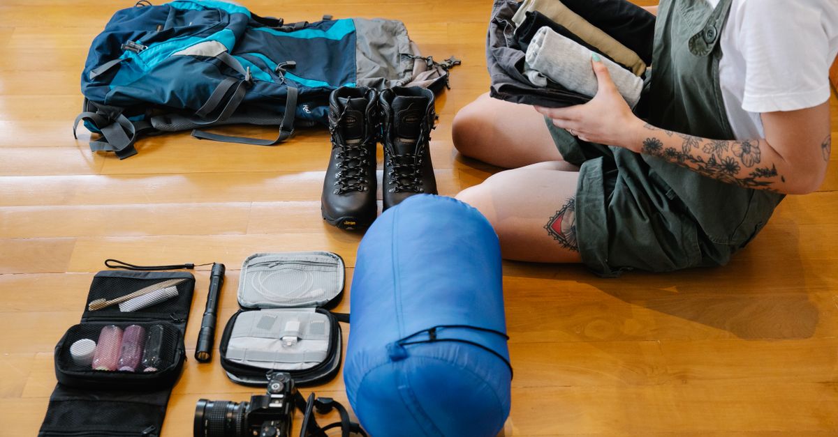 A person sits on a wooden floor packing a backpack, surrounded by hiking boots, a rolled sleeping bag, a camera, and a toiletry kit laid out beside them.