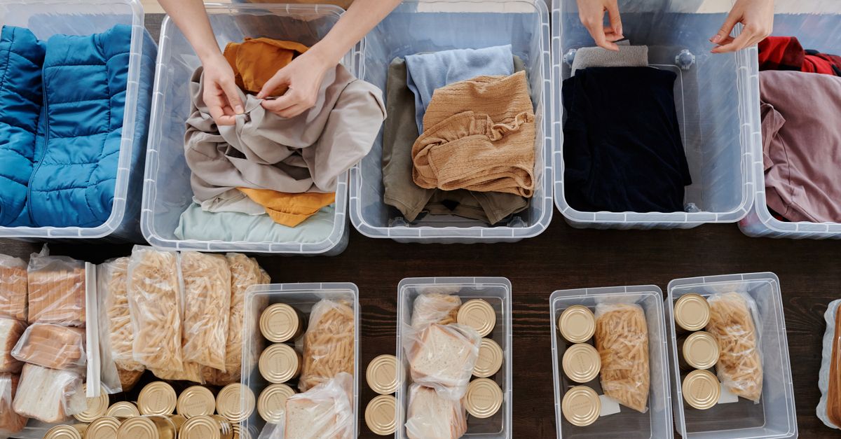 Overhead view of hands folding clothes into clear plastic storage bins arranged on a wooden floor alongside bins of packaged goods.