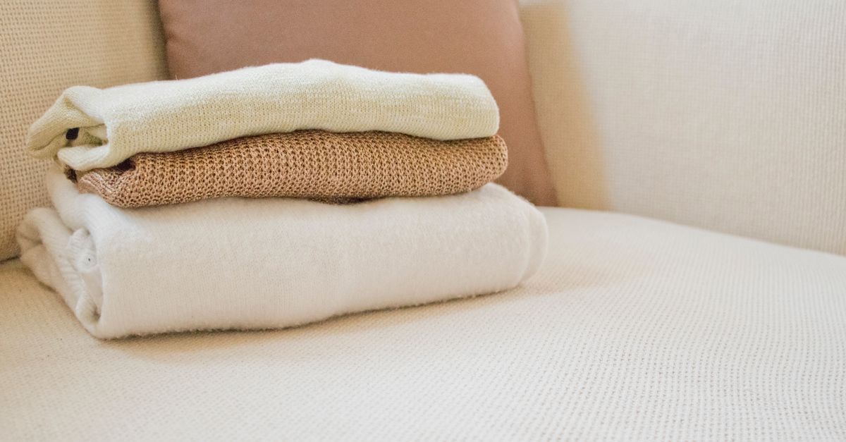 A neatly folded stack of a cream sweater, tan knit sweater, and white towel resting on a light-colored sofa.