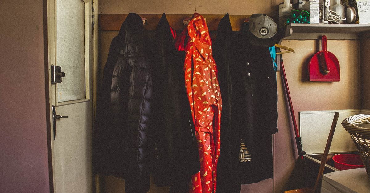 Coats including a dark puffer jacket hang on a wooden wall rack near a window in a home entryway, with household items on a shelf nearby.