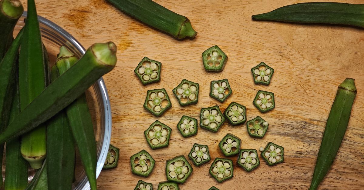 Sliced okra pieces scattered on a wooden cutting board alongside whole okra pods and a metal bowl of trimmed pods.