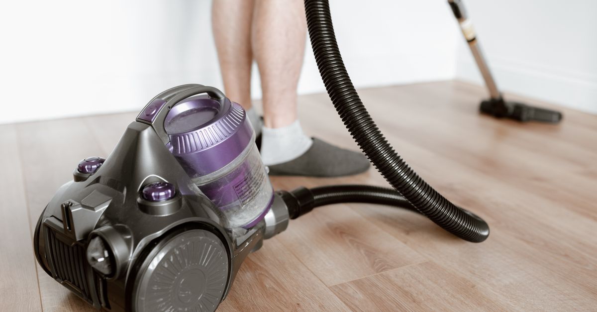 A canister vacuum cleaner with its hose and floor brush attached sits on a wooden floor next to a person's feet.
