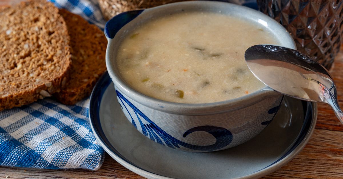 A creamy bowl of soup with a spoon rests on a plate beside slices of dark bread and a blue gingham cloth on a wooden table.