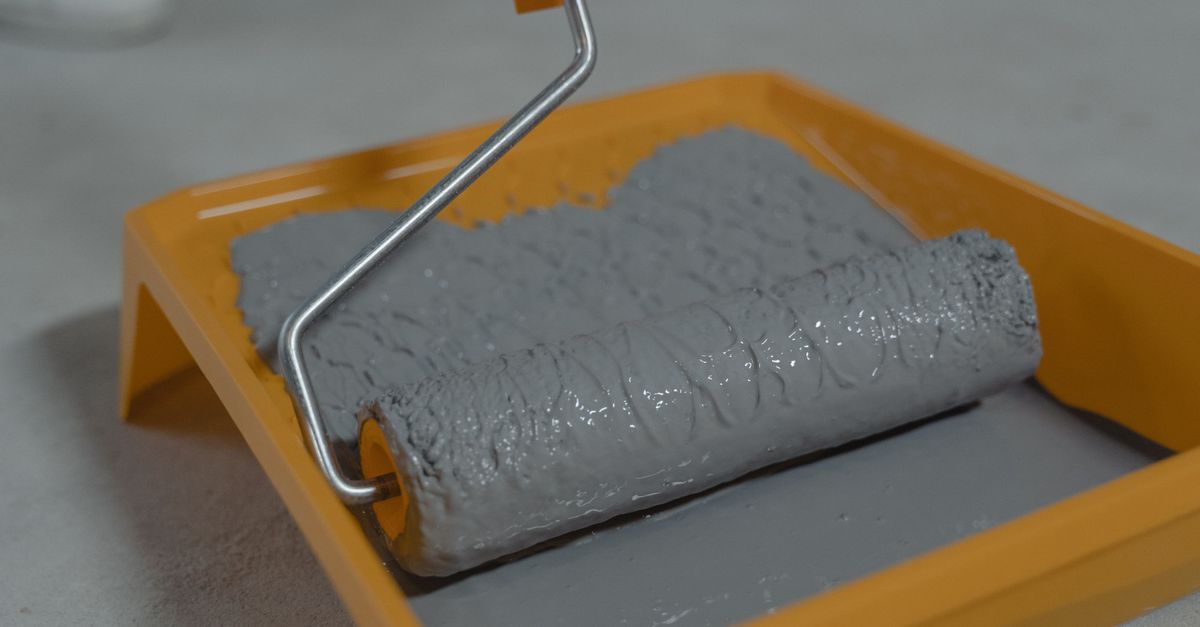A paint roller loaded with light gray paint resting in an orange plastic paint tray.