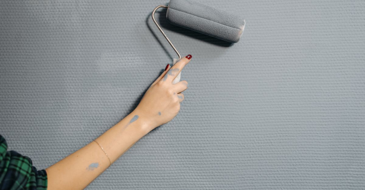 A hand with a paint roller applies gray paint to a wall, with a few drips visible on the arm.
