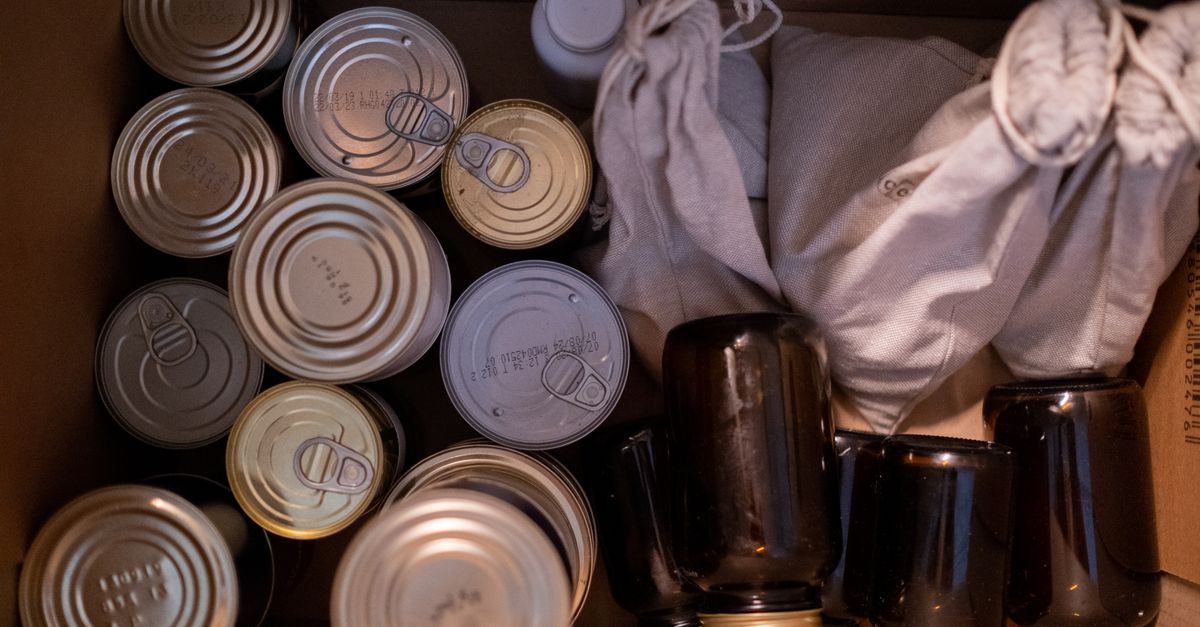 An overhead view of assorted canned goods and cloth-wrapped supplies packed into a cardboard box, arranged as emergency food storage.