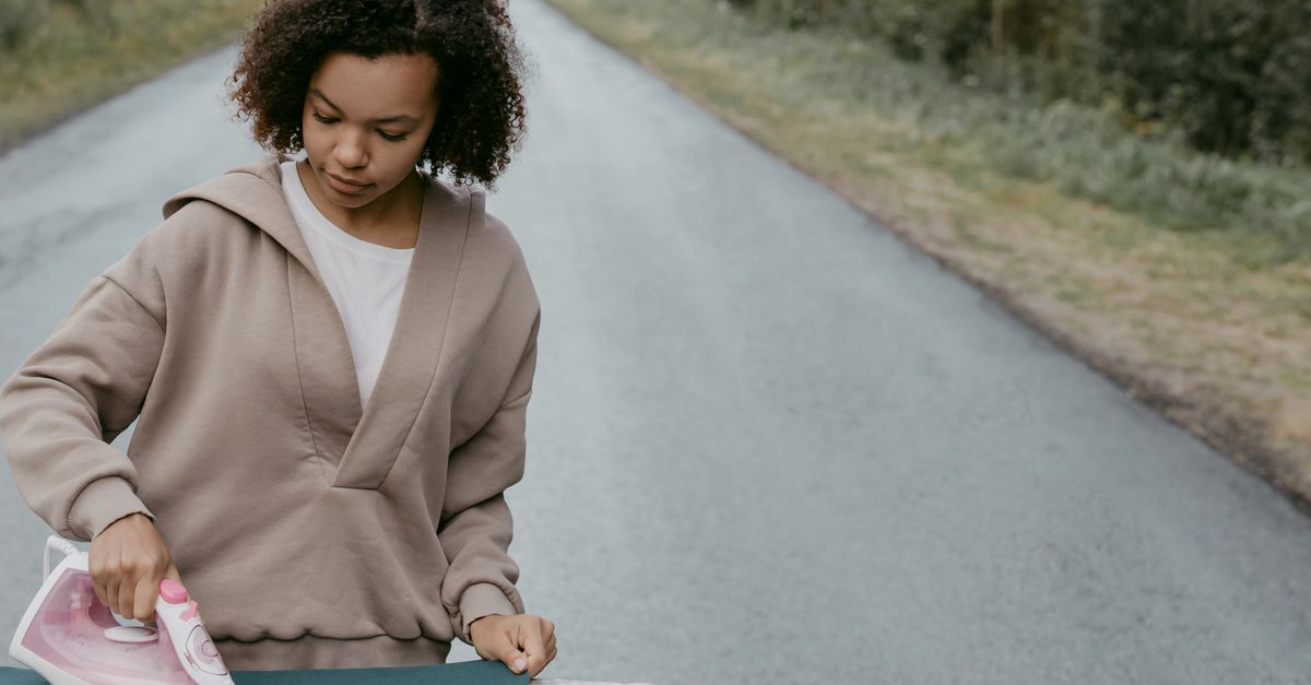 A woman irons a piece of fabric on an ironing board outdoors, with a road stretching into the distance behind her