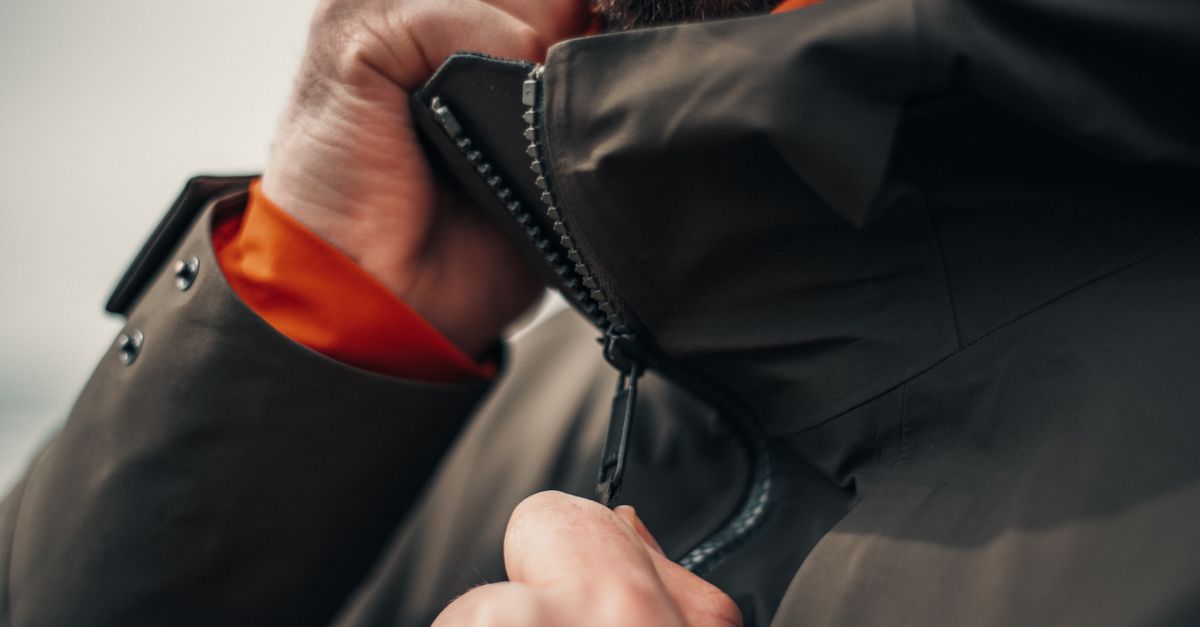 A close-up of a hand zipping up the collar of a dark technical outdoor jacket with an orange inner lining visible at the cuff.