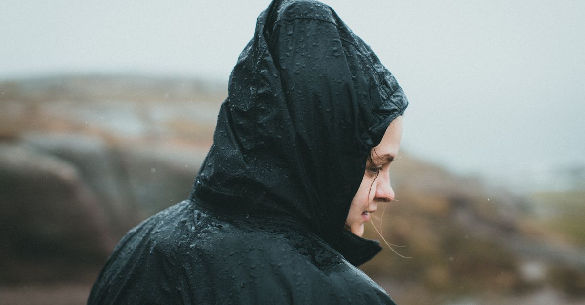 A person wearing a dark technical rain jacket with a hood, covered in beaded raindrops, seen from the side against a blurred outdoor background.