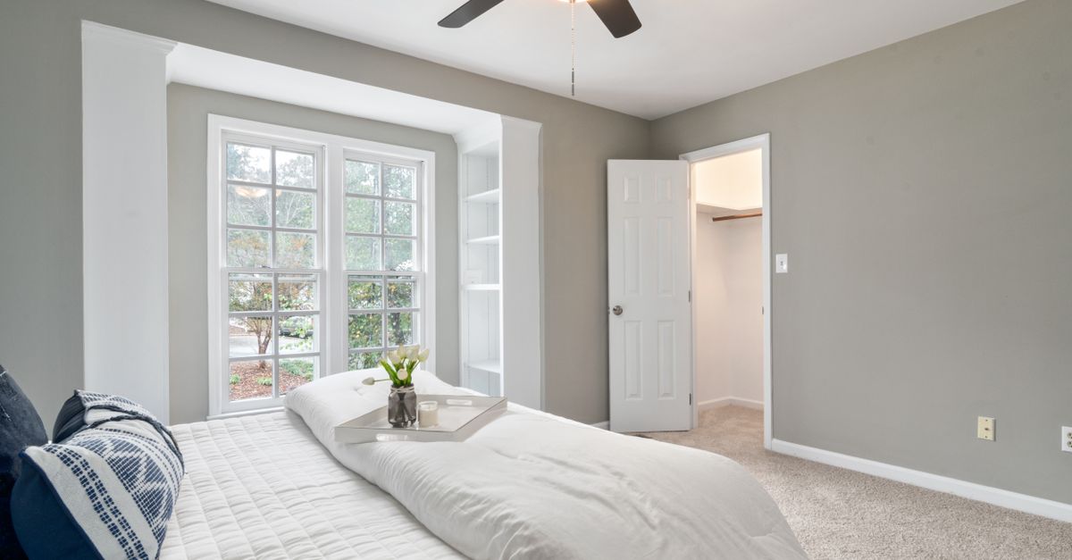 A bright bedroom with a large window framed in white, showing trees outside, morning light streaming in and a bed with white linens beneath it.
