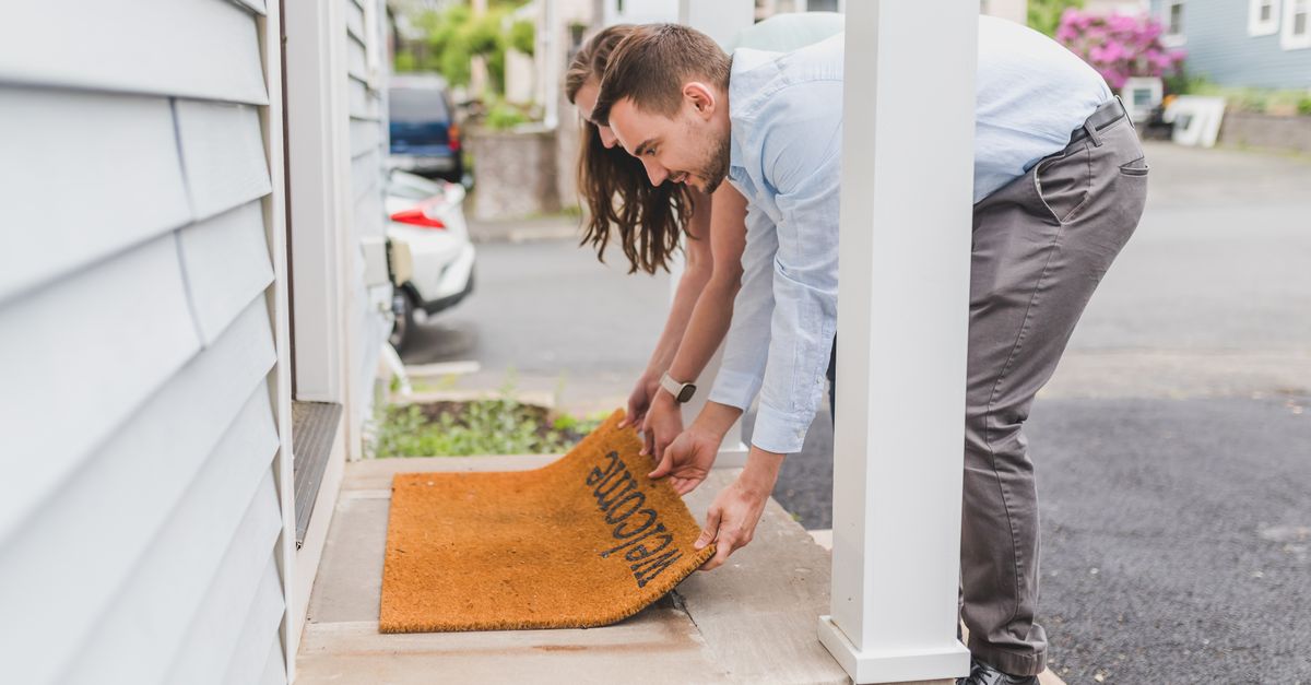 A man crouches on a porch step lifting the edge of a coir doormat printed with 'Welcome' lettering.