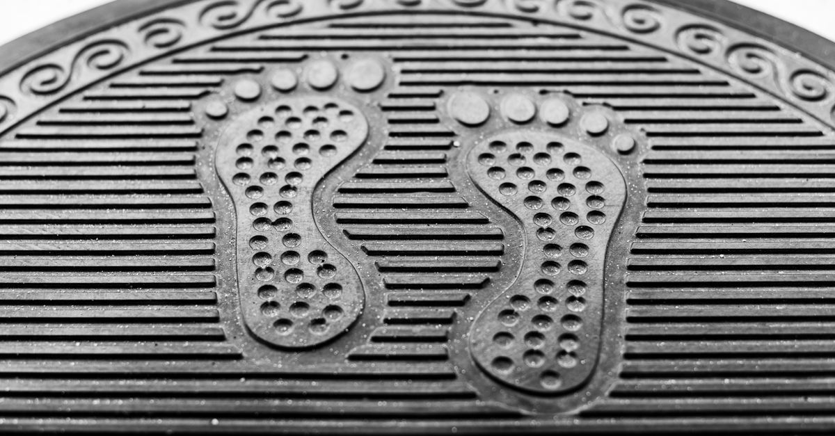 Close-up of a black rubber door mat with ribbed channel grooves and a decorative footprint design molded into the surface.