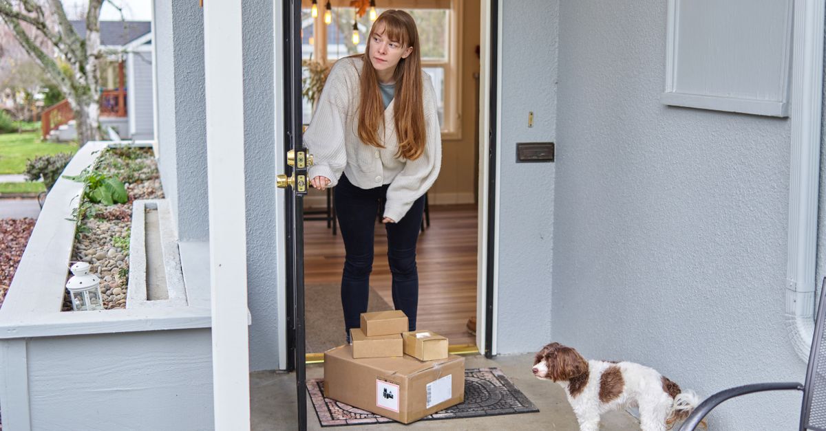 A doormat with cardboard packages sits at a front door as a woman opens the door and a dog waits nearby on the porch.