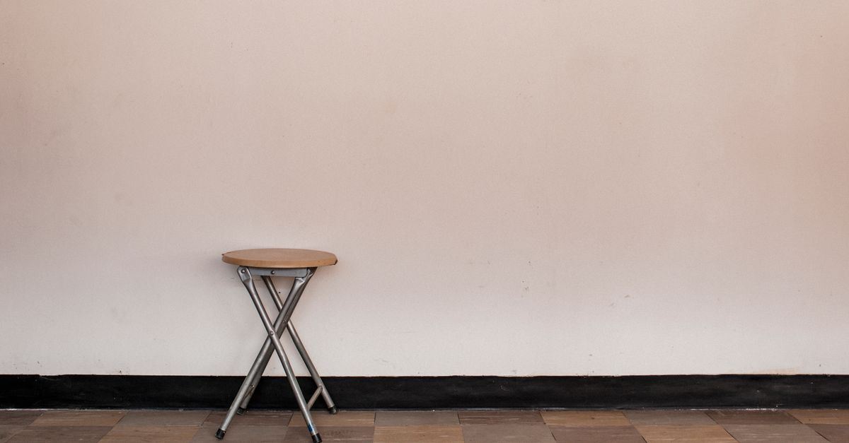 A small round folding stool with a wooden seat and silver metal legs stands against a plain wall.