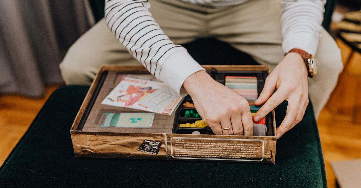A person's hands organizing colorful game pieces, cards, and tokens into the compartments of an open board game box.