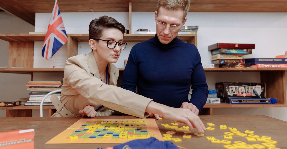 A man and woman sit at a table playing a board game with yellow tiles, with shelves of board games and books in the background.