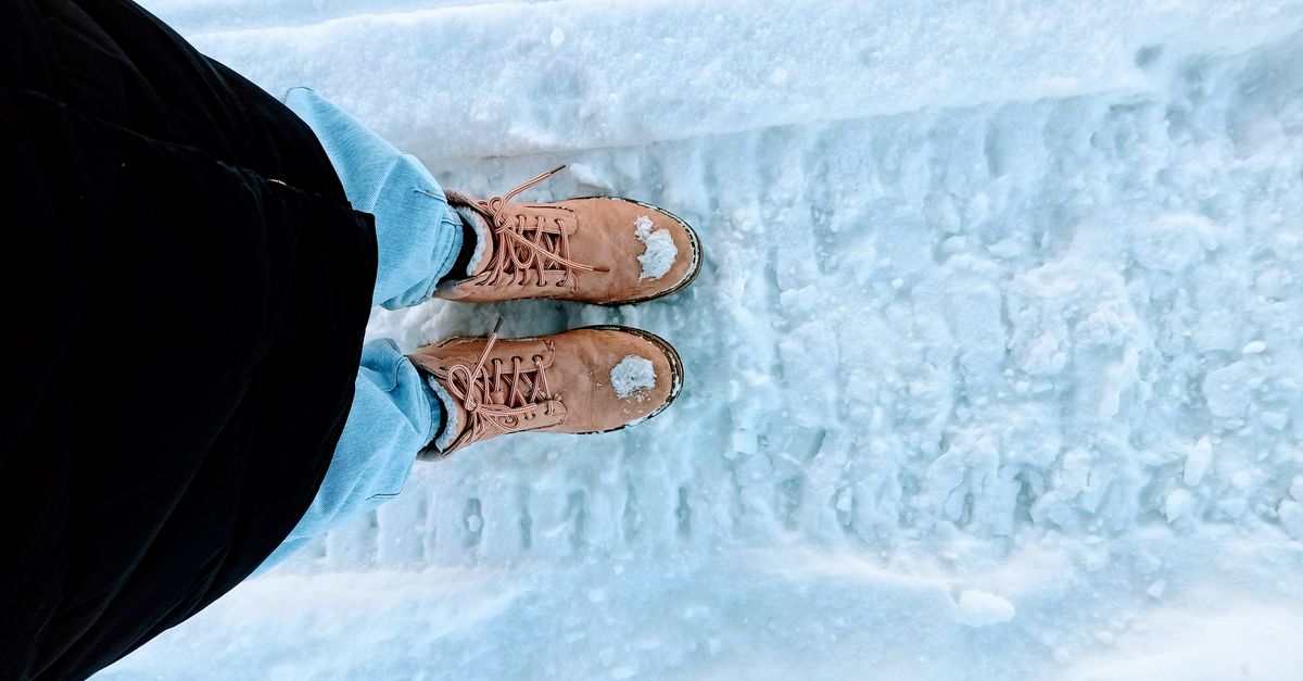 A person's tan lace-up winter boots covered in snow and slush marks while standing on an icy, tire-tracked path.