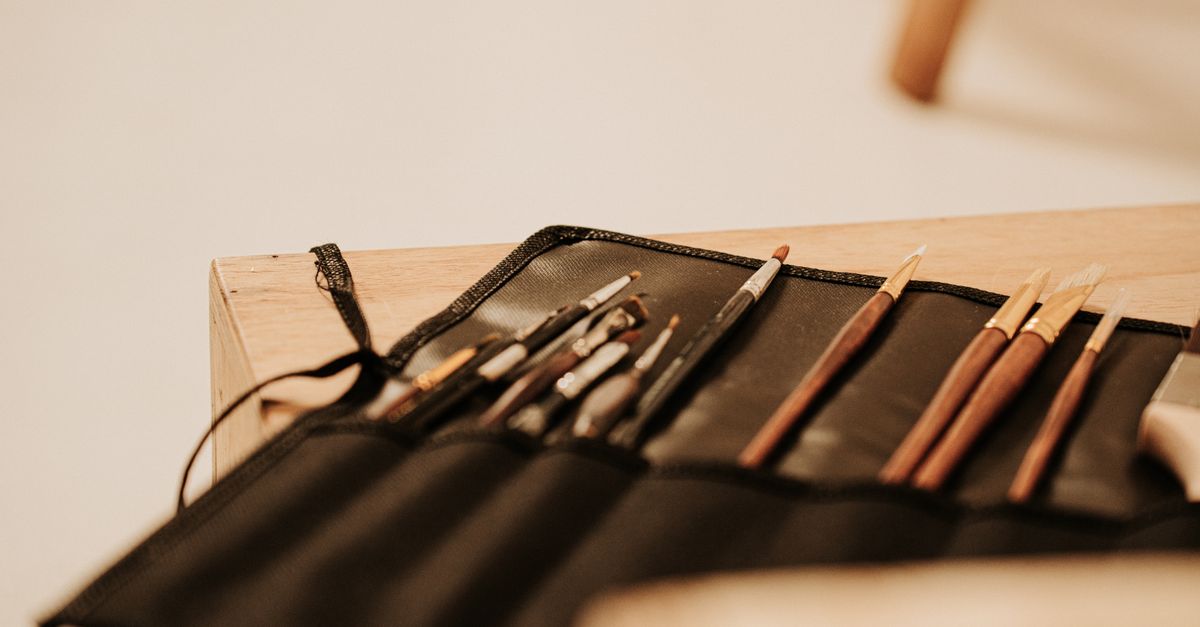 A canvas brush roll partially unrolled on a wooden surface, showing several artist brushes tucked into individual fabric sleeves.