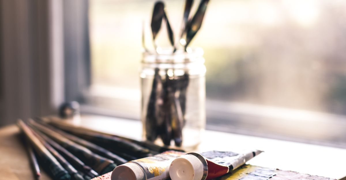 Rolled paint tubes lie beside a glass jar filled with paintbrushes near a window on a wooden surface.