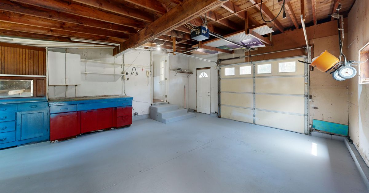 An empty garage workbench with blue and red cabinets, wooden shelving, bare walls, and a concrete floor seen from the front.