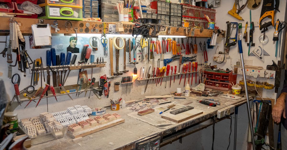 A cluttered garage workbench with pegboard walls holding pliers, screwdrivers, files, and small storage bins above a cement work surface covered with tools and materials.