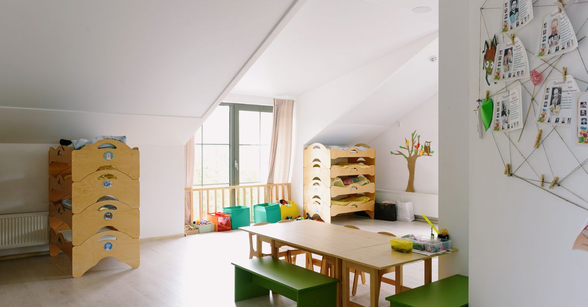 A children's room with low wooden shelving units, colorful storage bins on the floor, a wooden table with green benches, and a decorated tree on the wall.