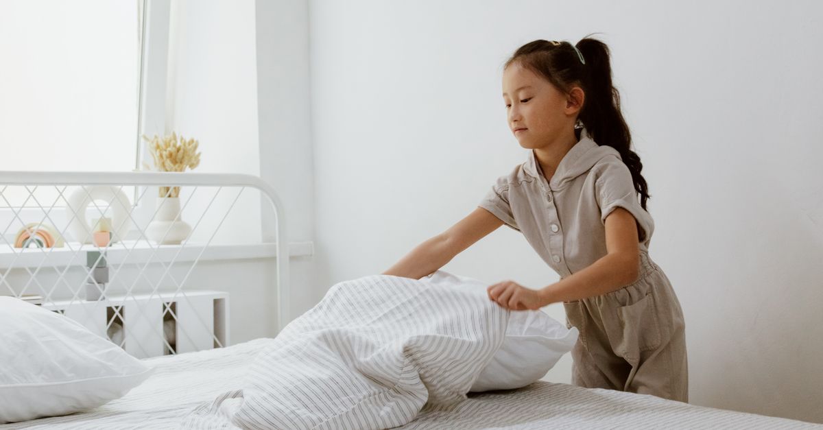 A young girl in a beige outfit tidies a striped blanket on a white bed frame in a bright, softly lit bedroom.