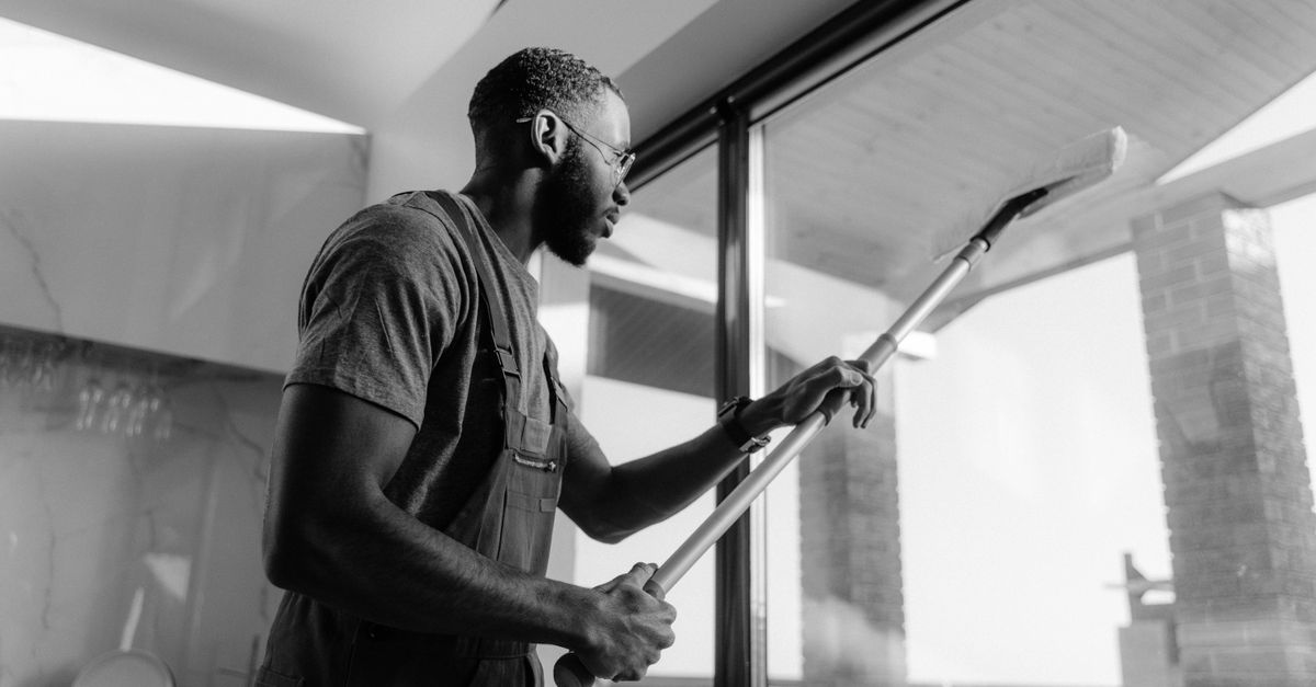 A man wearing overalls uses a long-handled squeegee to clean a large window in a black-and-white photo.