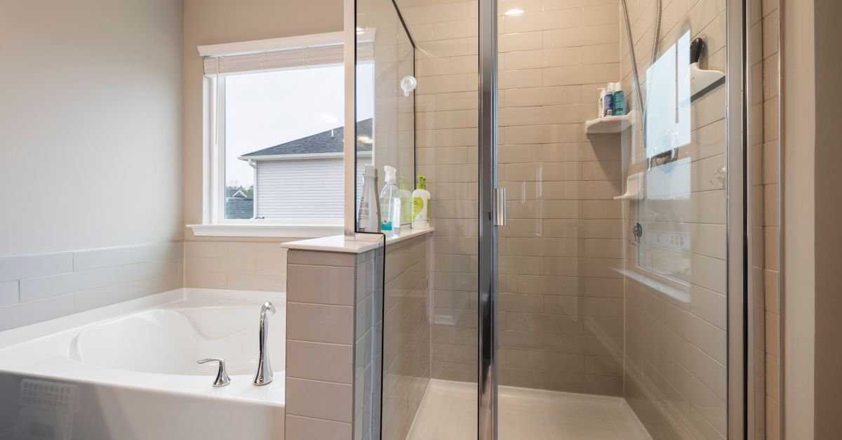 A clean glass shower door with chrome frame stands next to a bathtub in a bright bathroom, with a spray bottle placed on the tiled ledge.