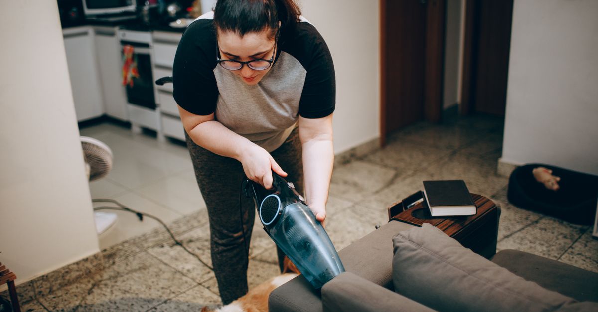 A woman vacuums a tiled floor near a couch with a small dog lying nearby, using a handheld vacuum cleaner.