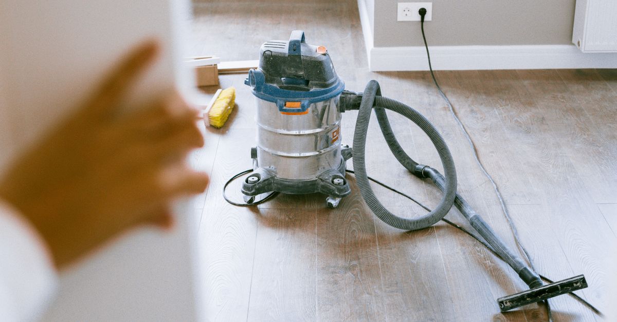 A canister vacuum cleaner with a hose and nozzle attachment sits on a hardwood floor as a shirtless man kneels beside it.