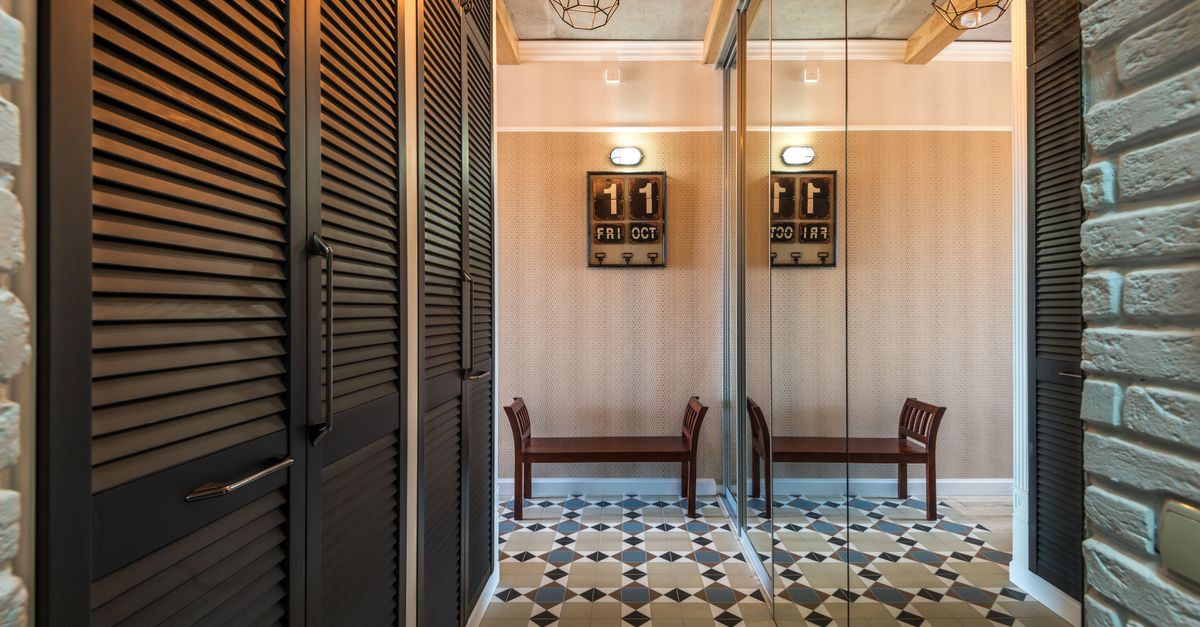 A dark wooden louvered closet with vertical slats stands in a minimalist entryway with patterned tile flooring and a mirrored wall.