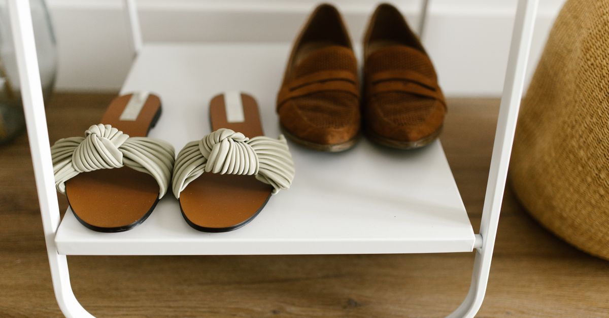 A white metal shoe rack shelf holding a pair of tan sandals and brown suede loafers near a wooden floor.
