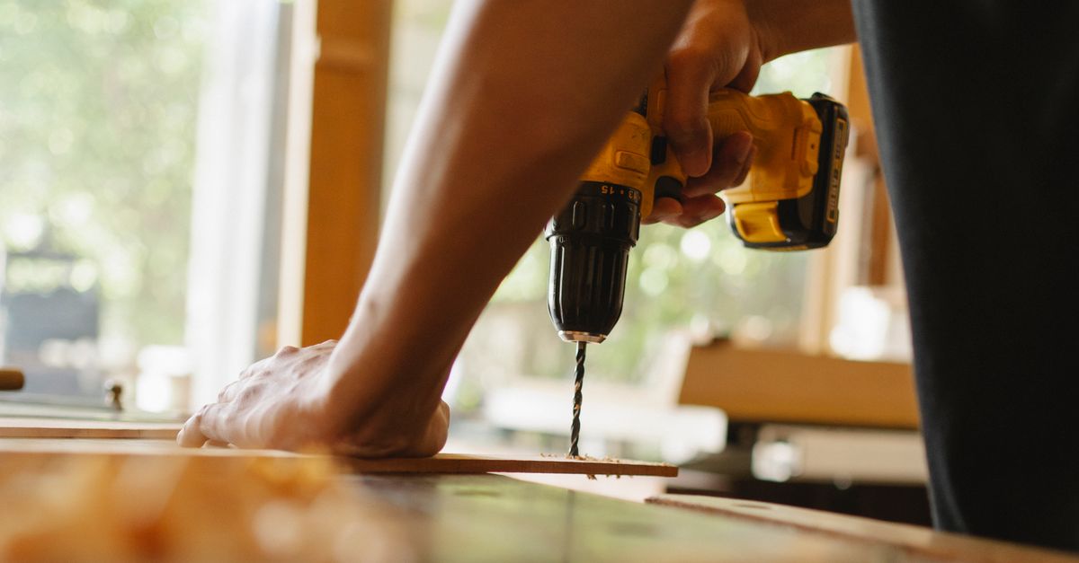A person drills into a piece of wood clamped to a workbench using a yellow and black power drill.