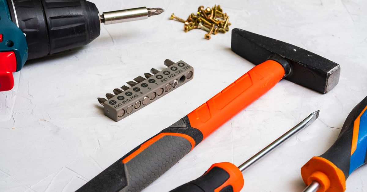A hammer, drill with driver bit, screws, and screwdrivers laid out on a light textured surface, with a driver bit set nearby.